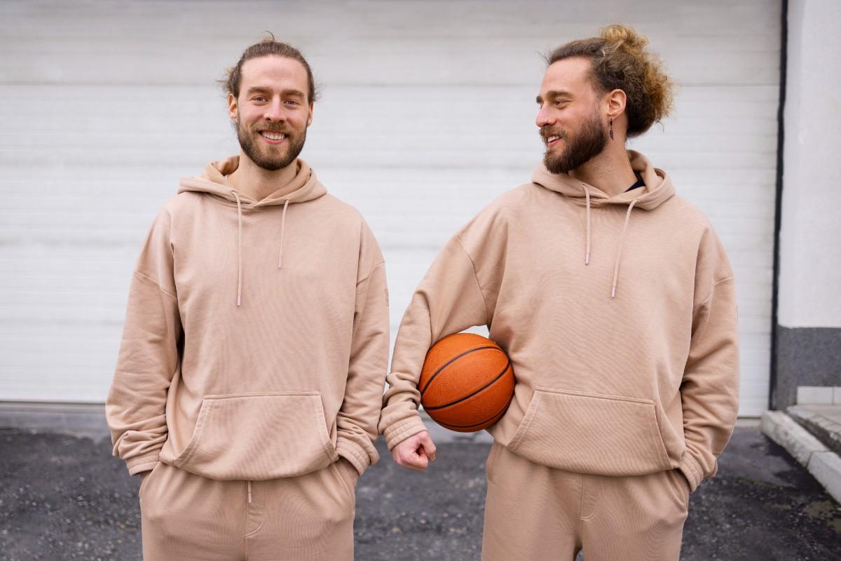 Two men wearing matching beige bulk sweatsuits, standing outdoors with one holding a basketball.